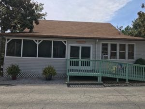 A single-story house with a brown roof and grey siding, featuring a screened-in porch on the left and windows on the right. A green ramp with a decorative railing and painted horse leads up to the entrance. Two potted plants are placed near the porch.