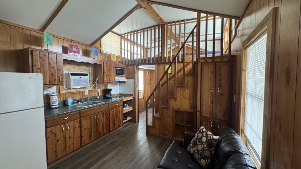 Interior of a cozy wooden cabin with a kitchen, staircase leading to a loft area, large windows, dark couch with pillows, and an abundance of natural wood throughout the walls and furniture.