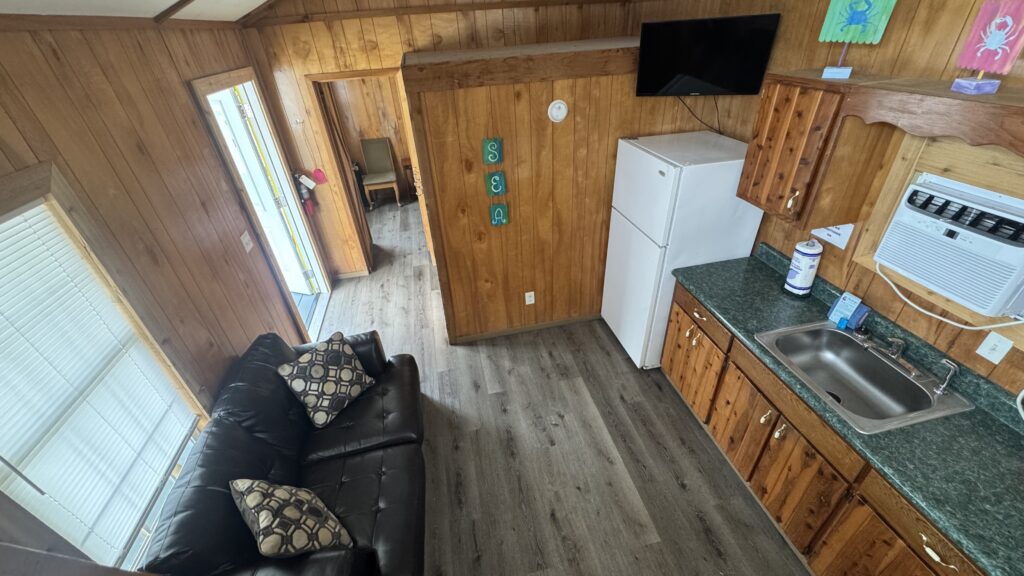 Interior of a small wooden cabin with a green countertop kitchen, white fridge, wall-mounted TV, black leather sofa with patterned pillows, and "SEA" decor on the wall. Light streams in from large windows.
