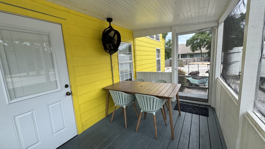 A screened porch with a yellow wall, white door, ceiling fan, wooden table, and four light blue chairs. Large windows show an outdoor view of trees, a fence, and neighboring houses.