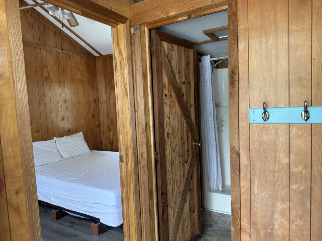 A small wooden cabin interior showing a bed with white linens in one room and an open door leading to a bathroom with a shower and a white curtain. There are wood-paneled walls and a rack with wall hooks.