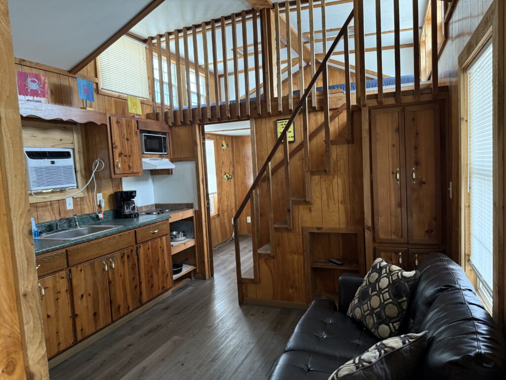 A cozy wooden cabin interior with a kitchenette, black couch, patterned pillows, stairs leading to a loft, and colorful banners hanging on the walls. Natural light comes through several windows.