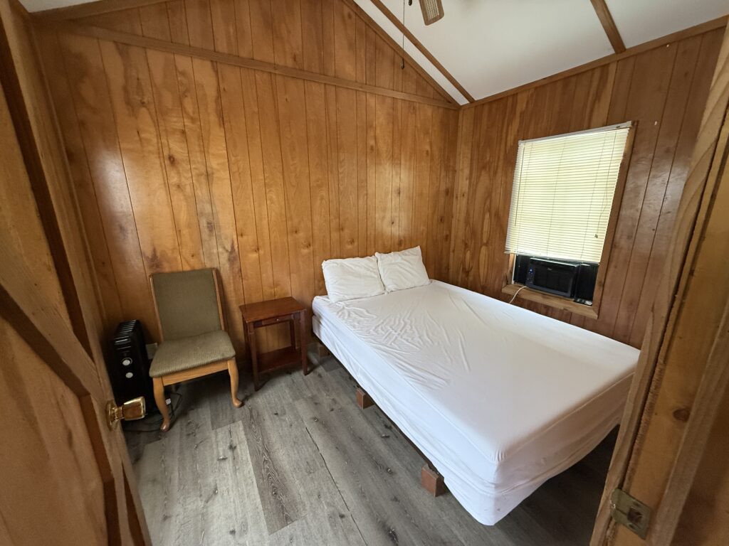 A small, simple bedroom with wood-paneled walls, a bed with white sheets and pillows, a wooden chair, a small side table, an air conditioner, and a window with closed blinds.