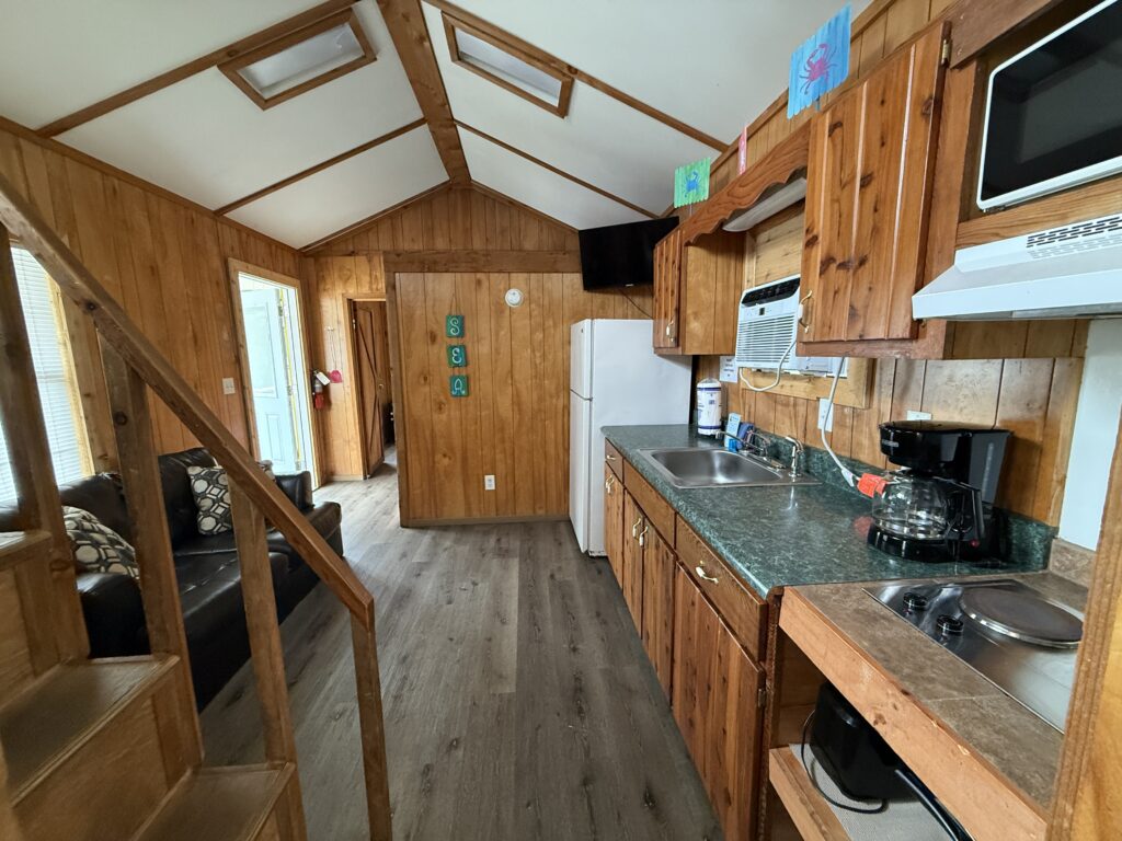 Interior of a cozy wooden cabin with a small kitchen, green countertop, appliances, stairs, a black couch, and bright natural light from windows. Colorful decorations are on the walls and counters.