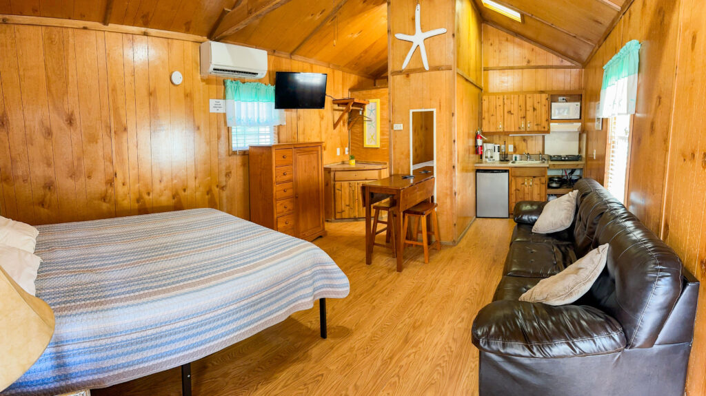 A cozy wooden cabin interior with a bed, black leather couch, small dining table, TV, dresser, and a kitchenette. The space has wood-paneled walls, vaulted ceiling, and natural light from small curtained windows.