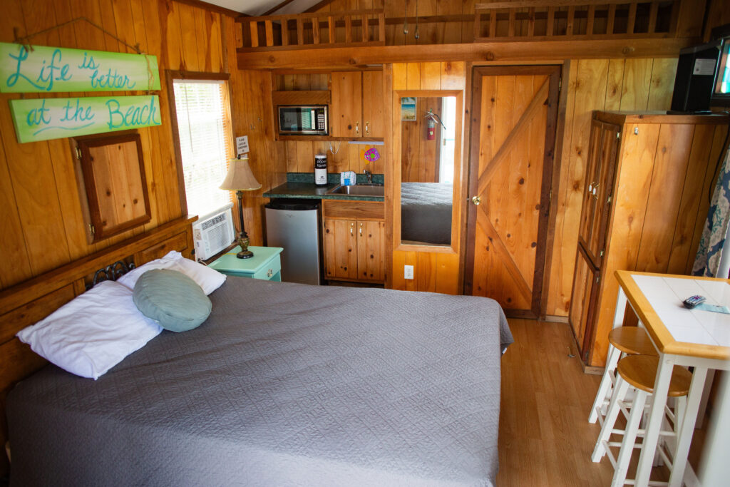 A cozy wooden cabin bedroom with a bed, gray quilt, small kitchenette, mirror, and a green sign on the wall reading "Life is better at the Beach." Natural light comes in through a window.