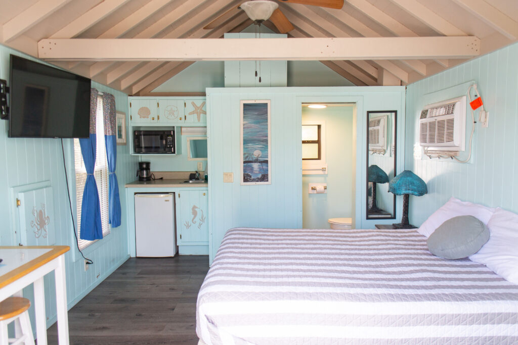 A cozy, light blue beach-themed cabin interior with a bed, small kitchenette, wall-mounted TV, and bathroom door. Decor includes sea life accents and blue lamps. A ceiling fan hangs above exposed beams.