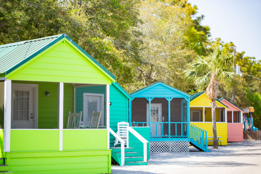A row of small, colorful beach cabins—painted green, blue, yellow, and pink—stand beside a gravel path under trees and a clear sky, with rocking chairs on some porches.