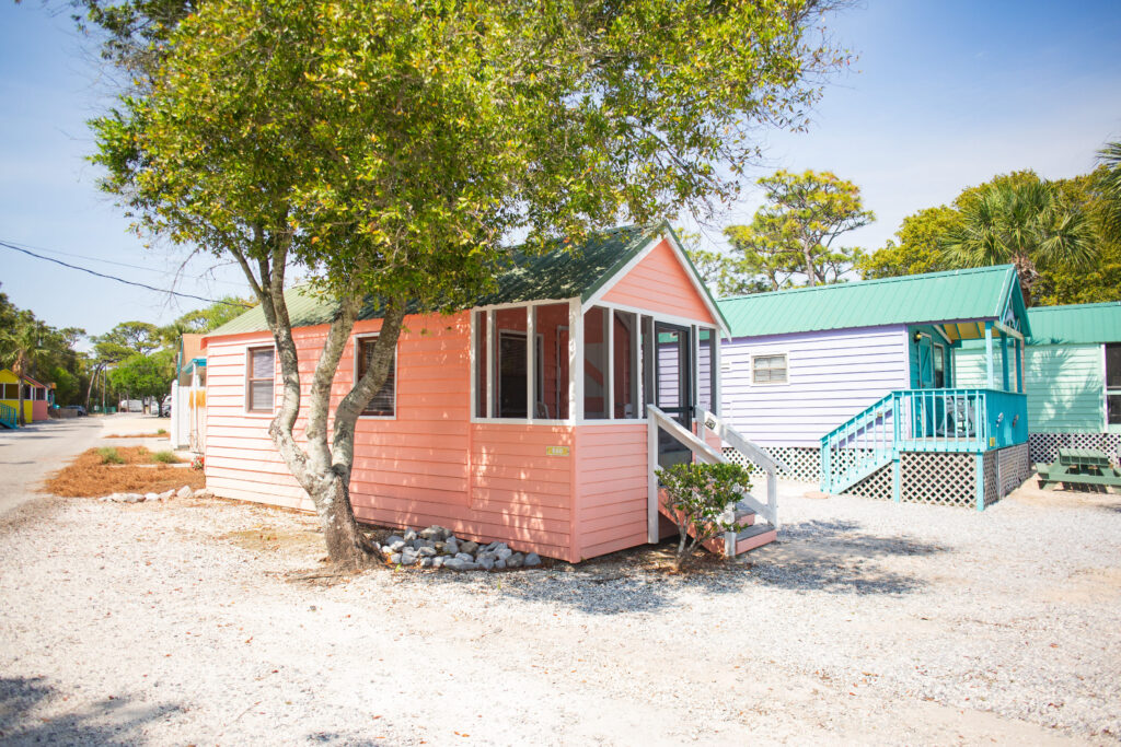 A small, pastel pink house with a green roof sits under a leafy tree on a gravel lot. Nearby, other colorful cottages with porches are visible under a bright blue sky.