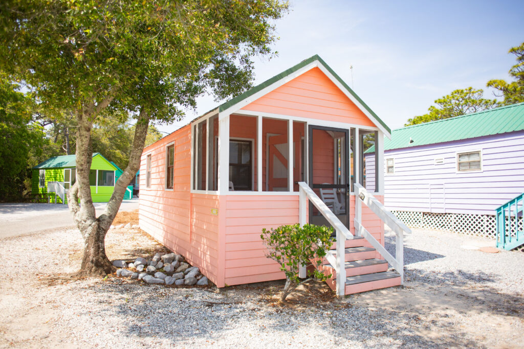 A small, pastel pink cottage with a screened porch sits under trees on a gravel lot. Nearby, another cottage painted green is visible, along with a turquoise-roofed building in the background.