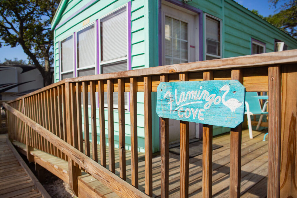 A turquoise house with white-trimmed windows has a wooden ramp leading to the entrance. A blue sign reading "Flamingo Cove" with a painted flamingo is attached to the railing.