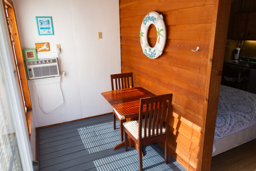 A small wooden dining table with two chairs sits by a window in a cozy room with wood-paneled walls, wall art, an air conditioner, and a decorative lifebuoy hanging above the table.