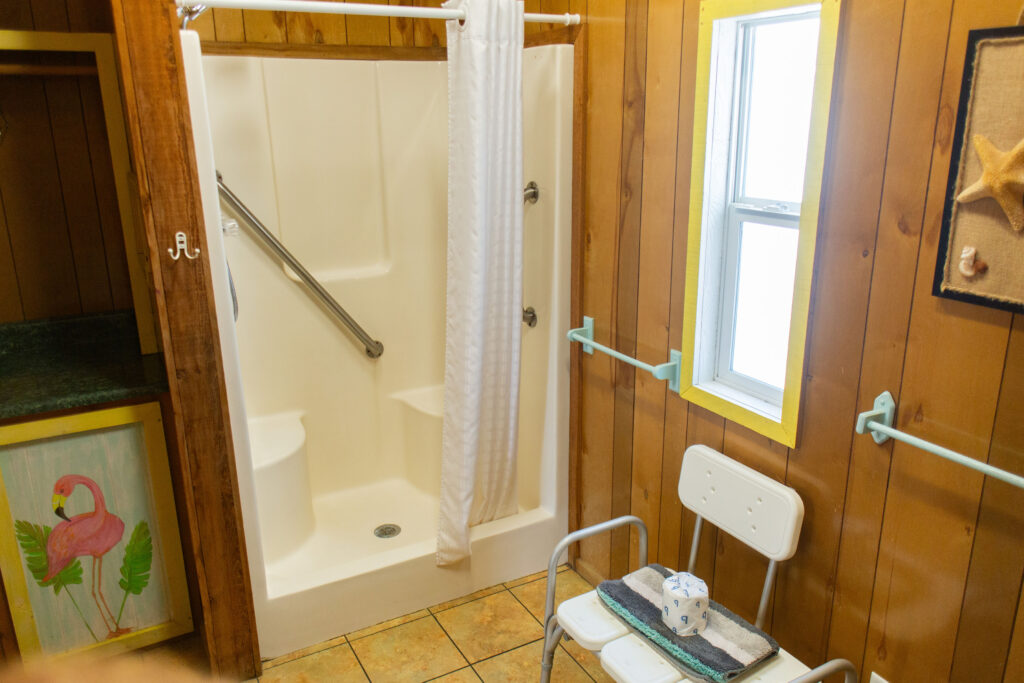 A small bathroom with wood-paneled walls, a white accessible shower with grab bars and a curtain, a white shower chair, a roll of toilet paper, and a window letting in natural light.