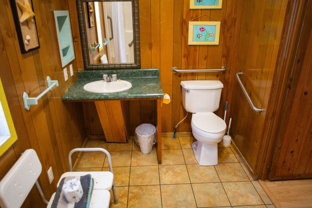 Accessible bathroom with a sink, toilet, grab bars, and a shower chair. The walls are wood-paneled, and there are framed pictures hanging above the toilet. The floor is tiled and a trash bin is under the sink.