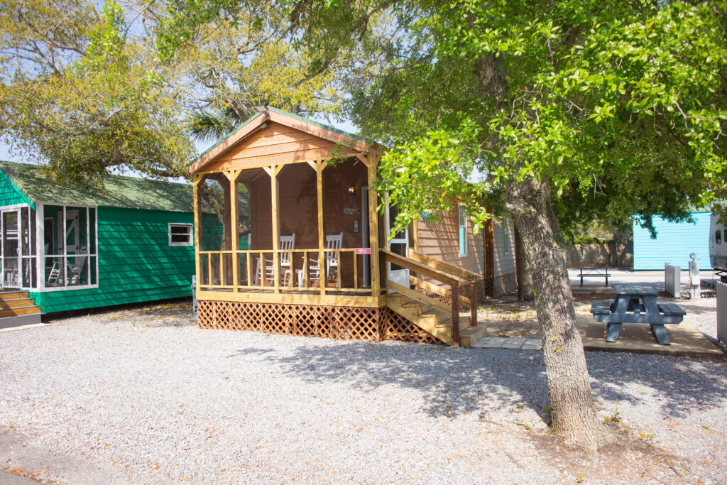 A small wooden cabin with a covered porch and two rocking chairs sits among trees. A picnic table is nearby, and another green cabin and a blue building are visible in the background.