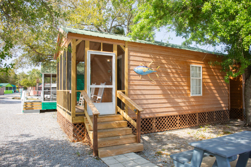 A small wooden cabin with a screened porch sits under leafy trees. There’s a fish decoration on the exterior wall, a picnic table in front, and gravel around the building. Sunlight creates dappled shadows on the cabin.