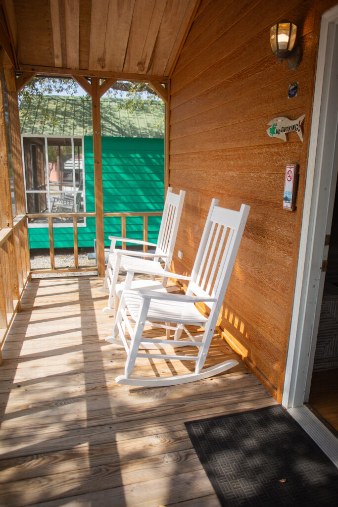 A wooden porch with two white rocking chairs facing outward, attached to a cabin. Sunlight casts shadows across the floor, and a small sign is mounted near the door. A green structure is visible in the background.