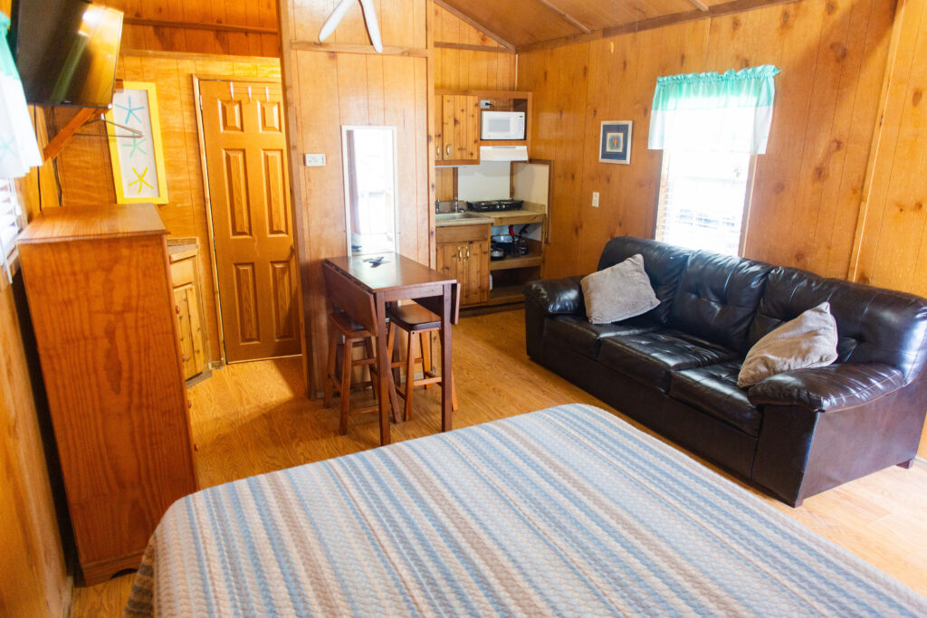 A cozy cabin interior with wood-paneled walls, a bed with a striped blanket in the foreground, a black leather sofa, a small kitchenette, a table with two chairs, and a door leading to another room. Natural light comes through a window.