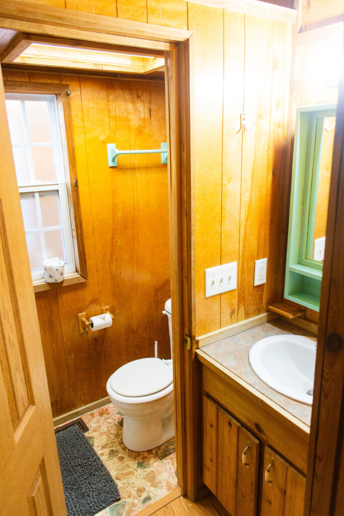 Small, rustic bathroom with wooden walls, a white toilet, a towel rack above the toilet, a window, a sink with a countertop, mirror, and cabinet, and a blue bath mat on a patterned floor.