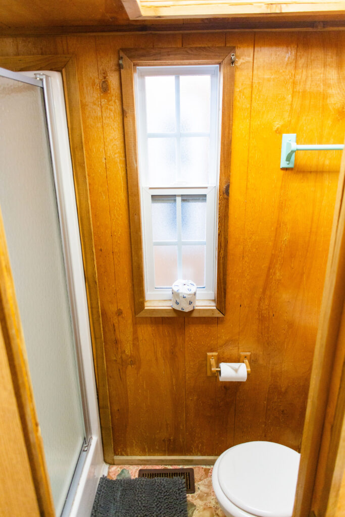 A small bathroom with wood-paneled walls, a shower with frosted glass door, a window, a towel rack, a toilet, a toilet paper holder, and a black floor mat. A toilet paper roll sits on the window ledge.
