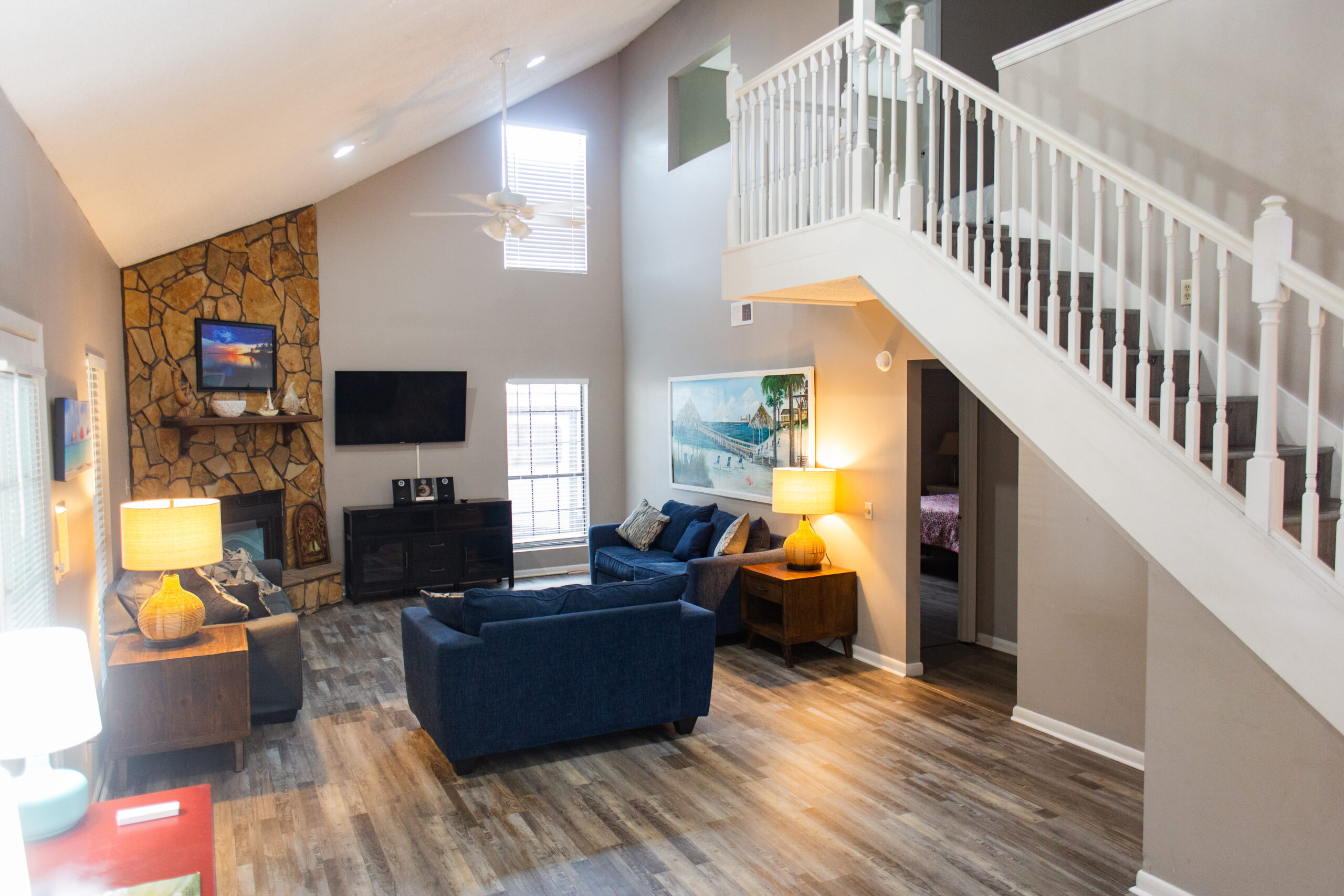 A modern living room with vaulted ceilings, blue sofas, a stone fireplace, TV, wooden flooring, and a white staircase leading to an upper level.