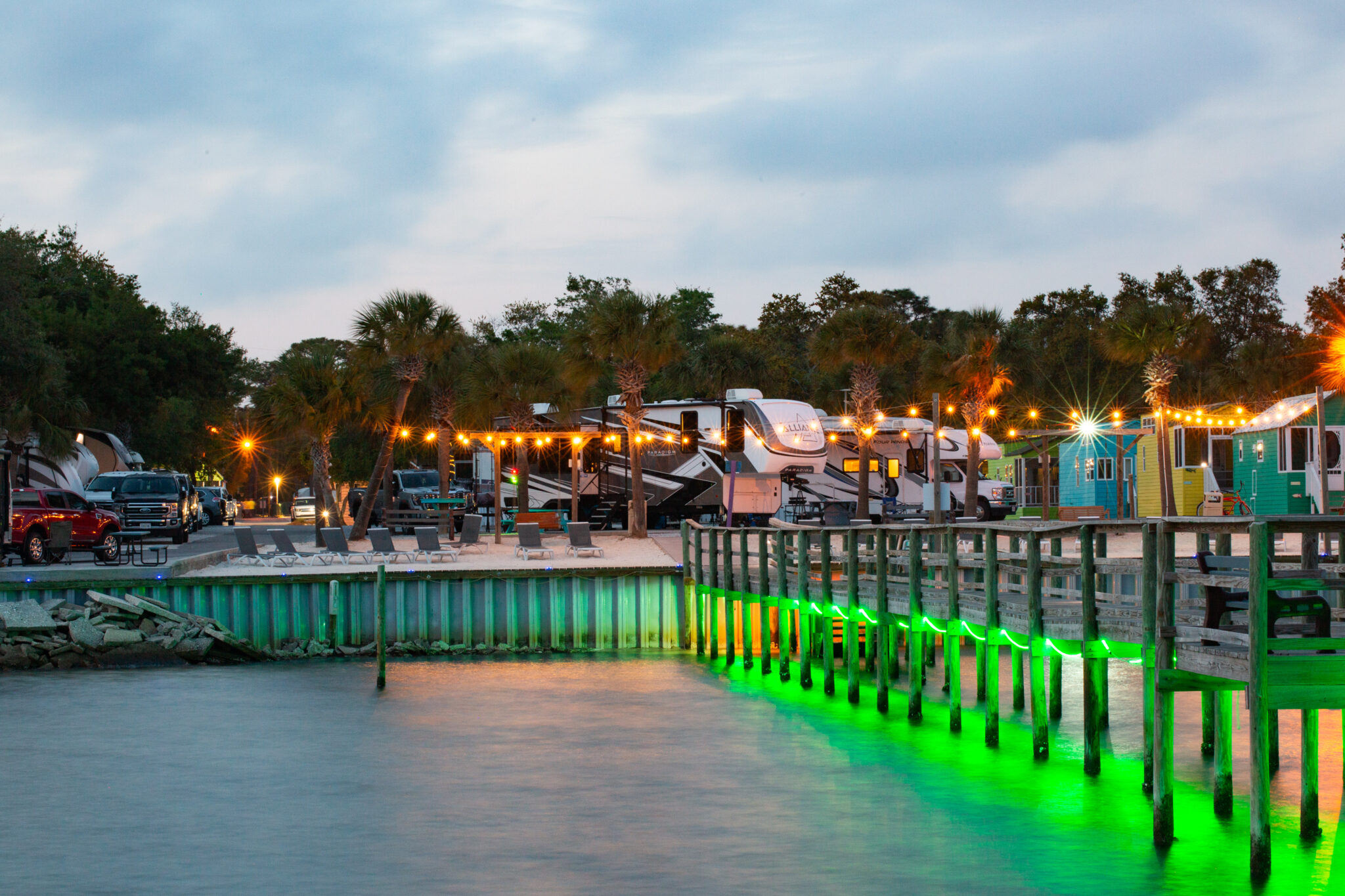 A waterfront RV park at dusk with string lights, palm trees, parked RVs, and a dock illuminated by green lights over the water.