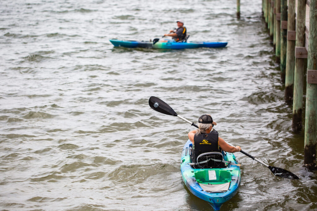 Two people in separate kayaks paddle on a choppy body of water near wooden docks, both wearing life jackets.