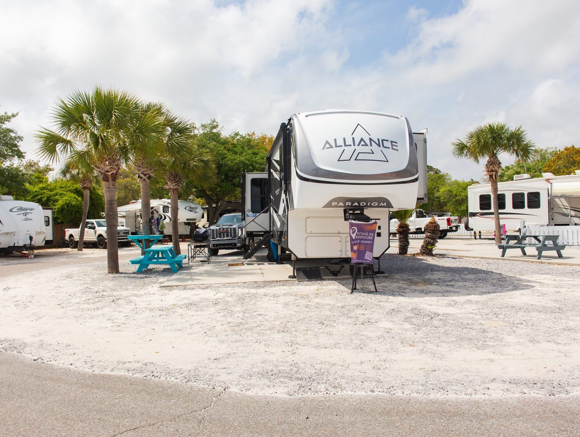 A large Alliance Paradigm RV is parked on a gravel site surrounded by palm trees, picnic tables, and other RVs under a partly cloudy sky.