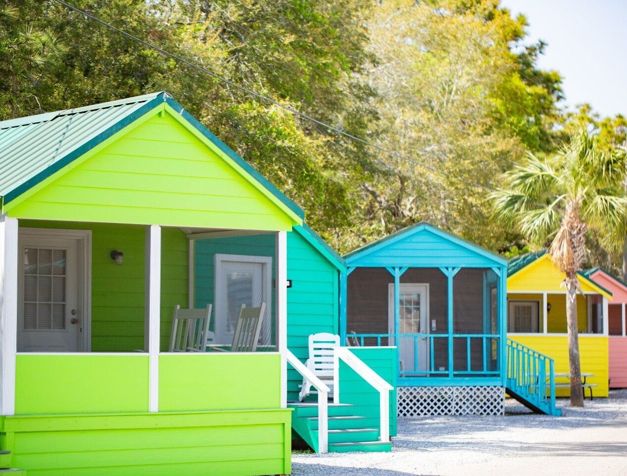 Row of small, brightly colored beach cabins with front porches, set against trees and clear sky.