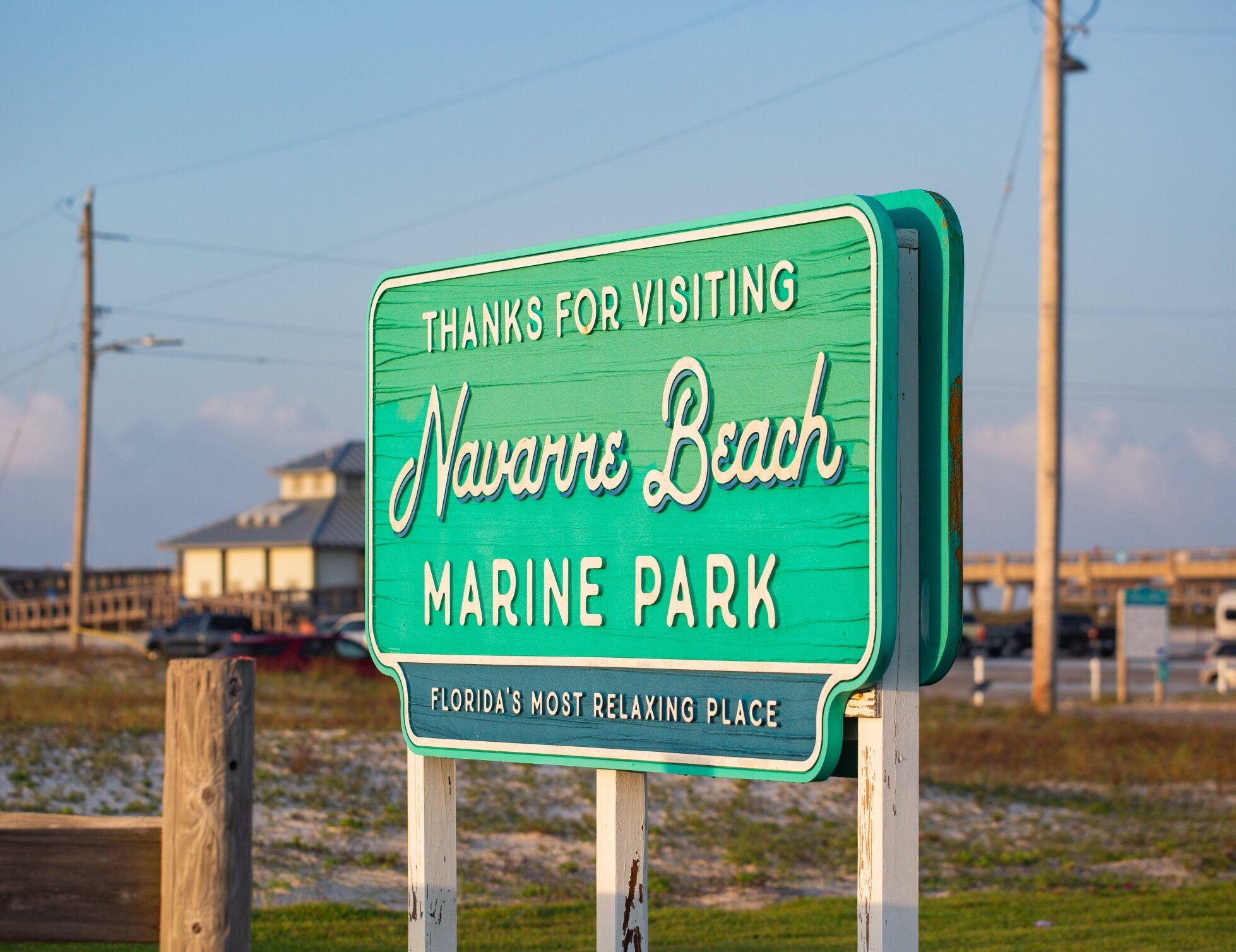 A green sign reads "Thanks for visiting Navarre Beach Marine Park, Florida's most relaxing place," with a beach, buildings, and utility poles in the background.