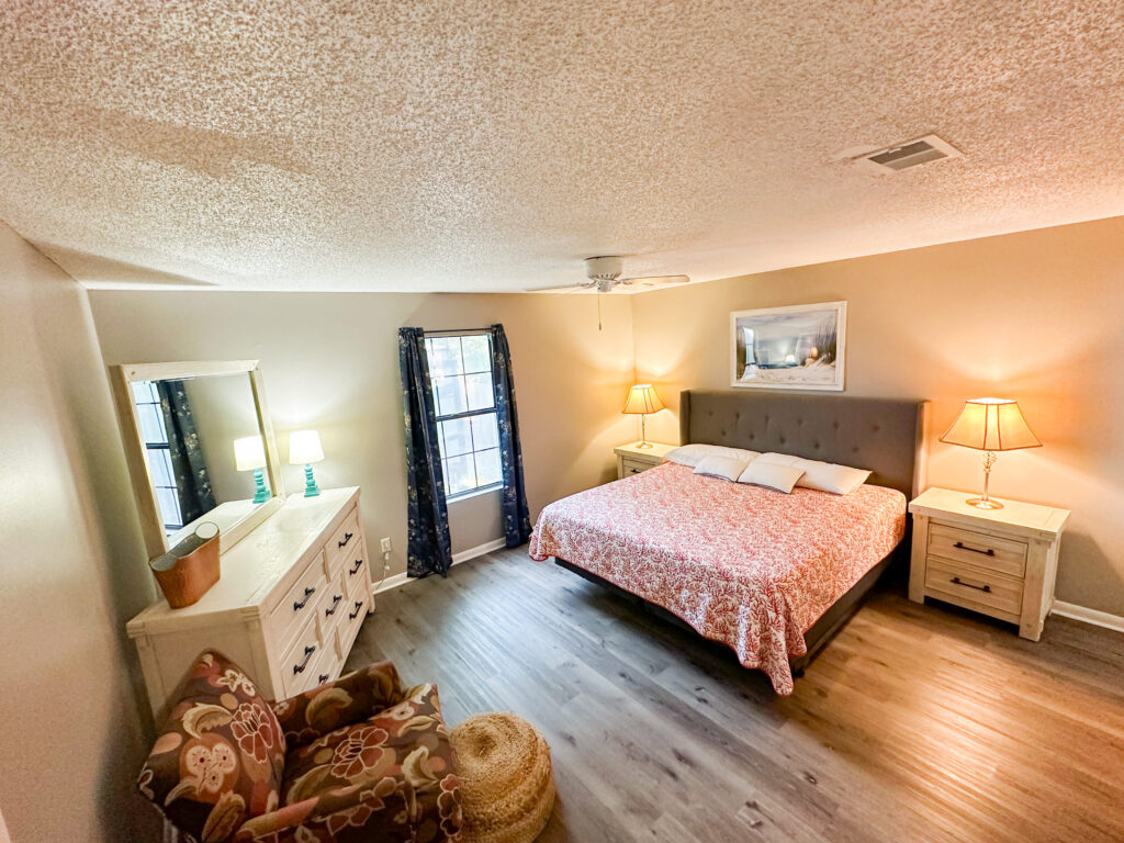A bedroom with a king-size bed, two nightstands with lamps, a dresser with a mirror, an armchair, and two windows with curtains.