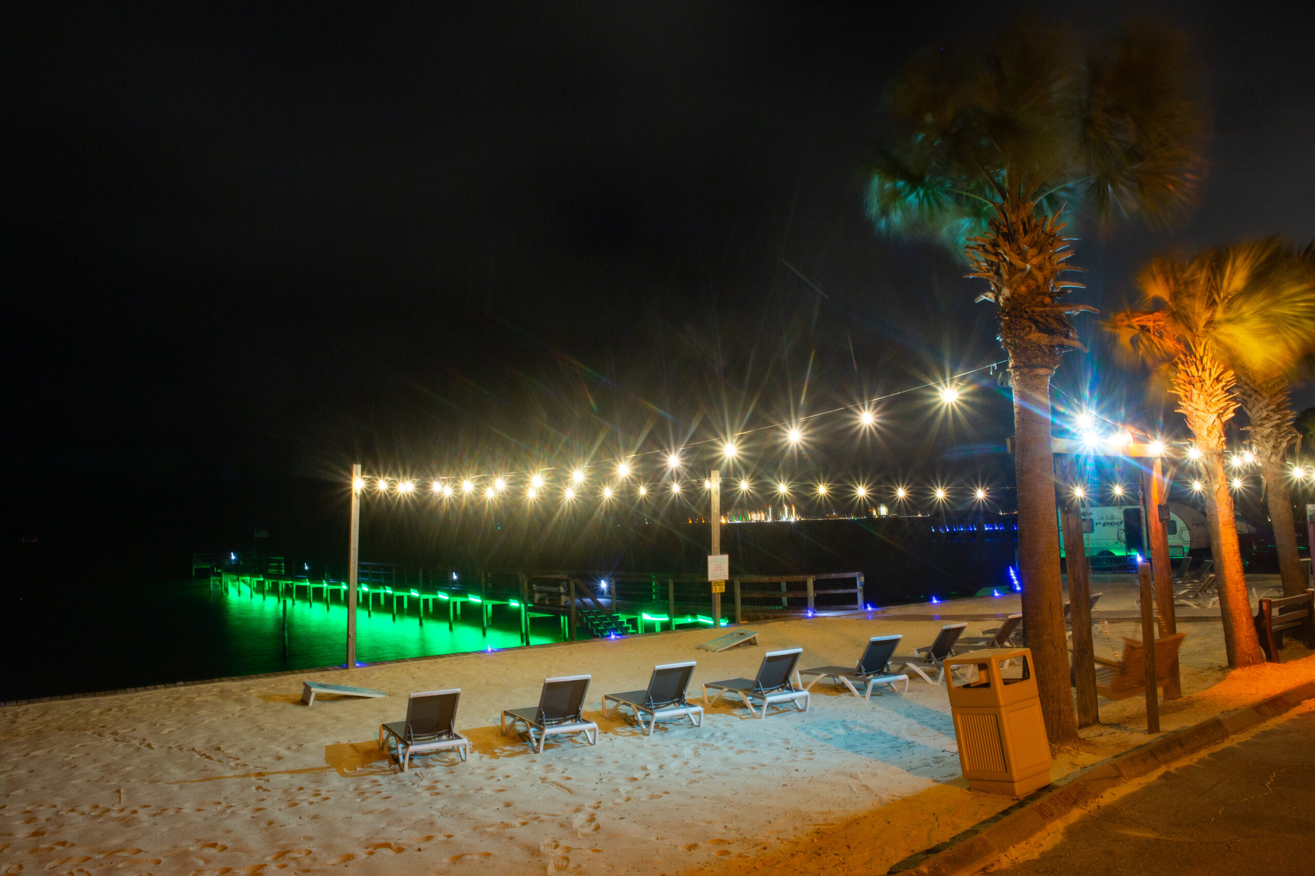 A nighttime beach scene with empty lounge chairs, palm trees, string lights overhead, and a pier lit with green lights over the water.