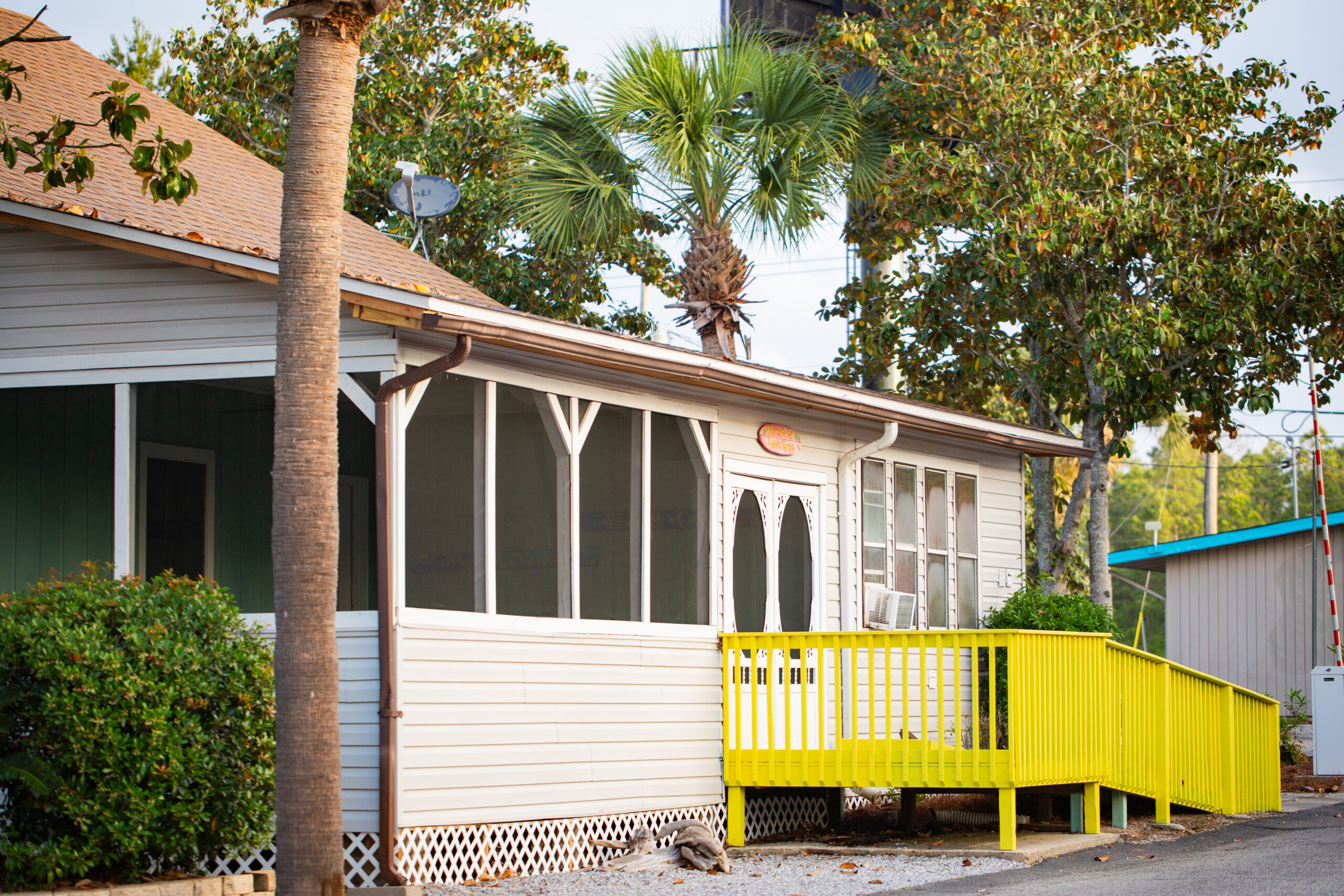 A single-story building with a screened porch and a bright yellow ramp, surrounded by palm trees and greenery.