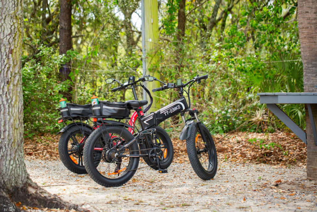 Two electric bikes with water bottles attached are parked on a gravel path in a wooded outdoor setting.