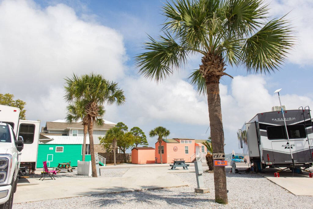 RVs and colorful tiny houses are parked among palm trees in a campground under a partly cloudy sky.