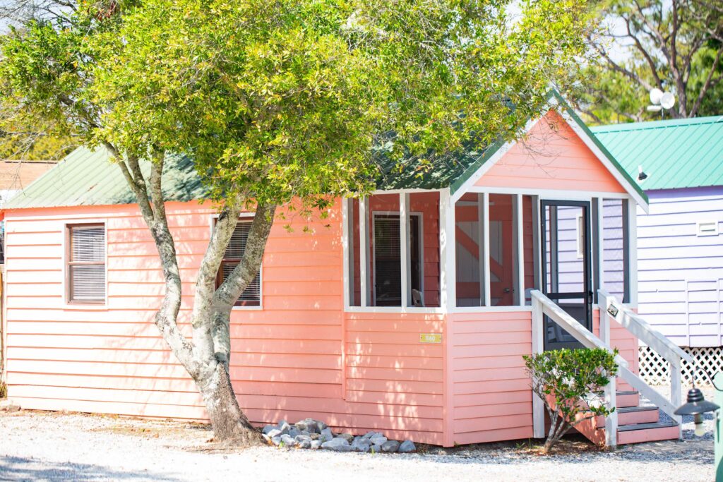 A small, peach-colored wooden house with a green roof and screened porch sits under a tree on a sunny day.