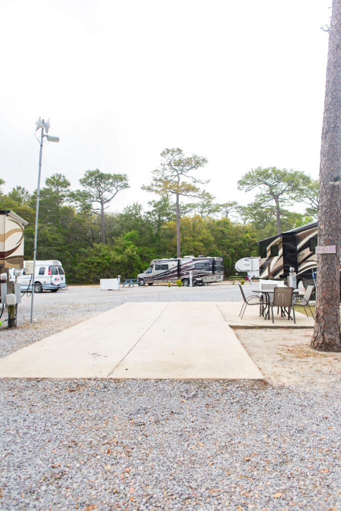 A gravel RV park with empty parking pad in the foreground, RVs parked in the background, trees, and outdoor seating on the right side.
