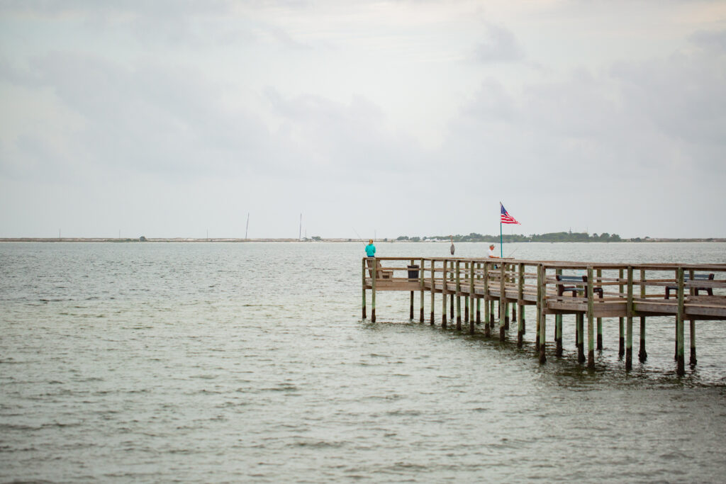 A person stands on a wooden pier over the water, near an American flag, with a cloudy sky and distant land visible in the background.