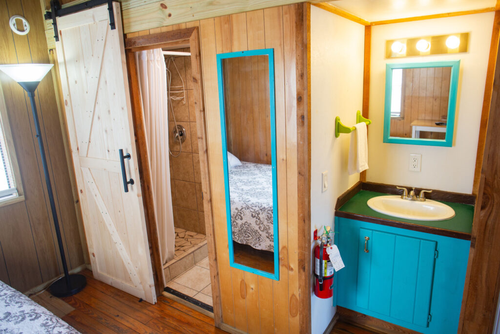 A small bathroom with a turquoise vanity, white sink, wall mirror, and towel rack. An adjacent shower with a sliding barn door is visible, along with part of a bedroom.