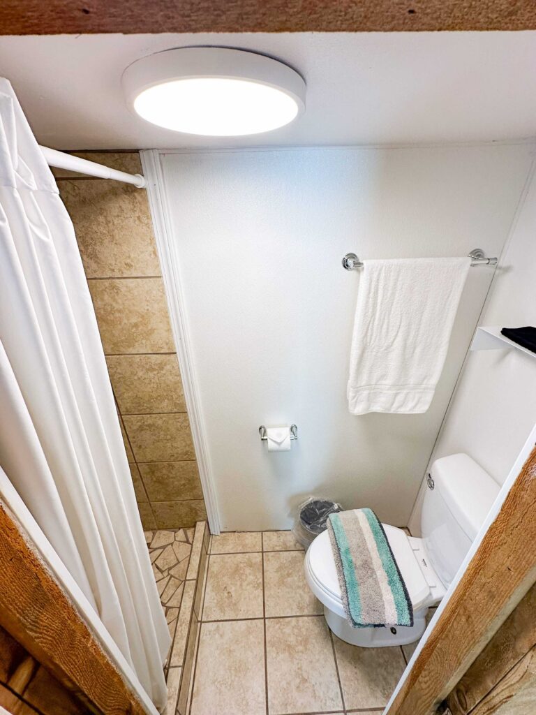 Small bathroom with a shower, toilet, towel hanging on a rack, toilet paper holder, trash can, and a striped towel on the toilet lid under a ceiling light.