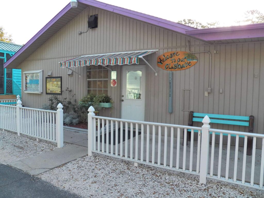 A small, light brown clubhouse with a striped awning, white fence, teal bench, and a sign reading "Welcome to our Clubhouse" near the entrance.