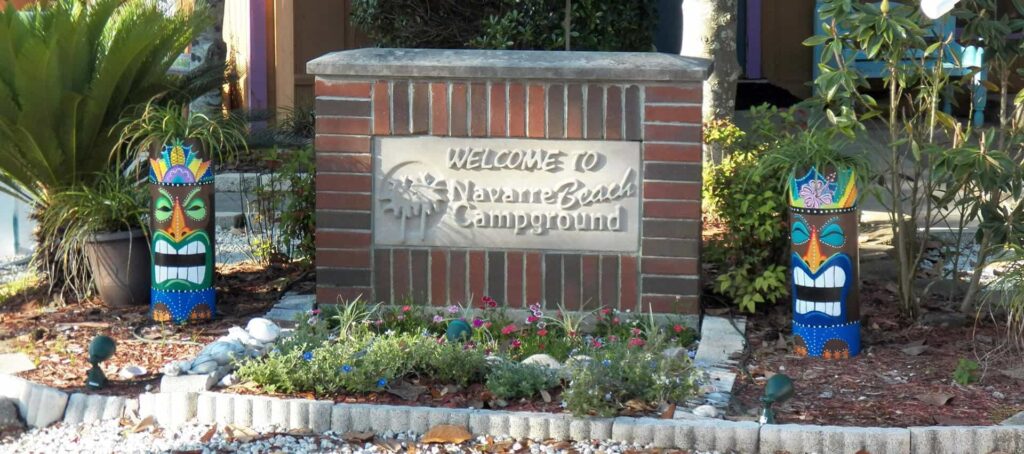Brick sign reading "Welcome to Navarre Beach Campground," surrounded by plants, flowers, and two colorful tiki statues on either side.