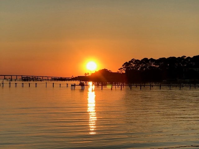 Sunset over calm water with silhouetted trees and docks in the distance; the sun reflects off the water's surface.