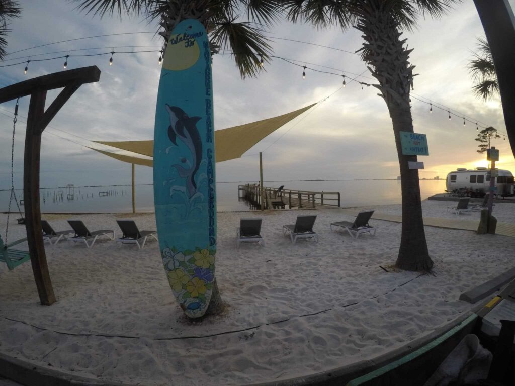 A sandy beach with lounge chairs, a surfboard with a dolphin design, palm trees, string lights, and a wooden dock extending into the water at sunset.