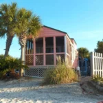 A small pink beach house with a screened porch is surrounded by palm trees, sand, and a white picket fence under a clear blue sky.