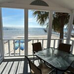 A glass-top table with four chairs sits on a screened porch overlooking a sandy beach, kayaks, palm tree, and ocean under a clear blue sky.