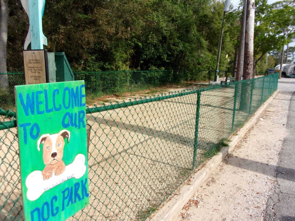 A fenced-in dog park area with a sign that reads "Welcome to our Dog Park" featuring a cartoon illustration of a dog; trees and a path are visible in the background.