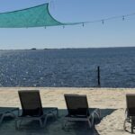 Four lounge chairs on a sandy beach face a calm body of water, shaded by a teal canopy, with a wooden pier and string lights in the background.