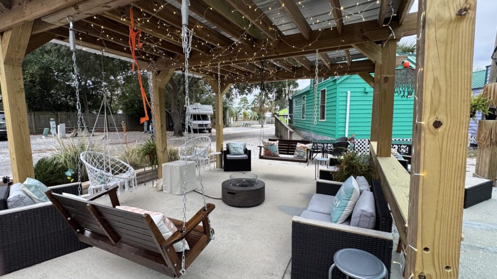 Outdoor seating area under a wooden pergola with string lights, swing benches, wicker chairs, a fire pit, and a turquoise building in the background.