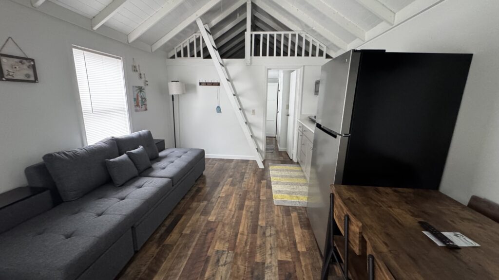 Small living area with a gray sofa, wooden floor, dining table, refrigerator, and a loft accessed by a ladder. White walls and ceiling with exposed beams. Natural light from a window.